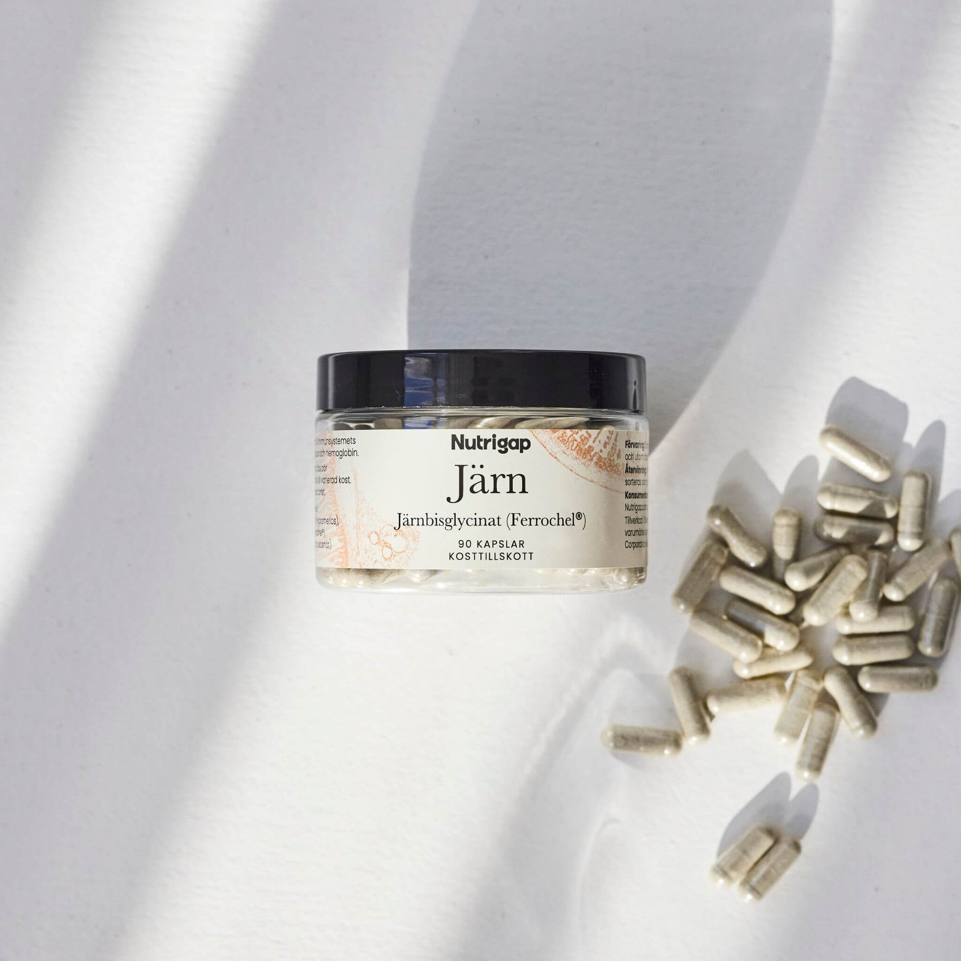 Burk med järntillskott i solljus med kapslar bredvid på vit bakgrund